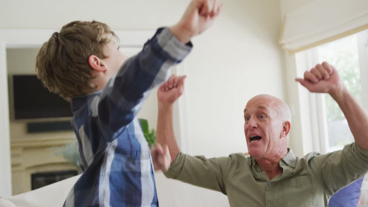 Happy caucasian grandfather and grandson jumping and raising hands in living room