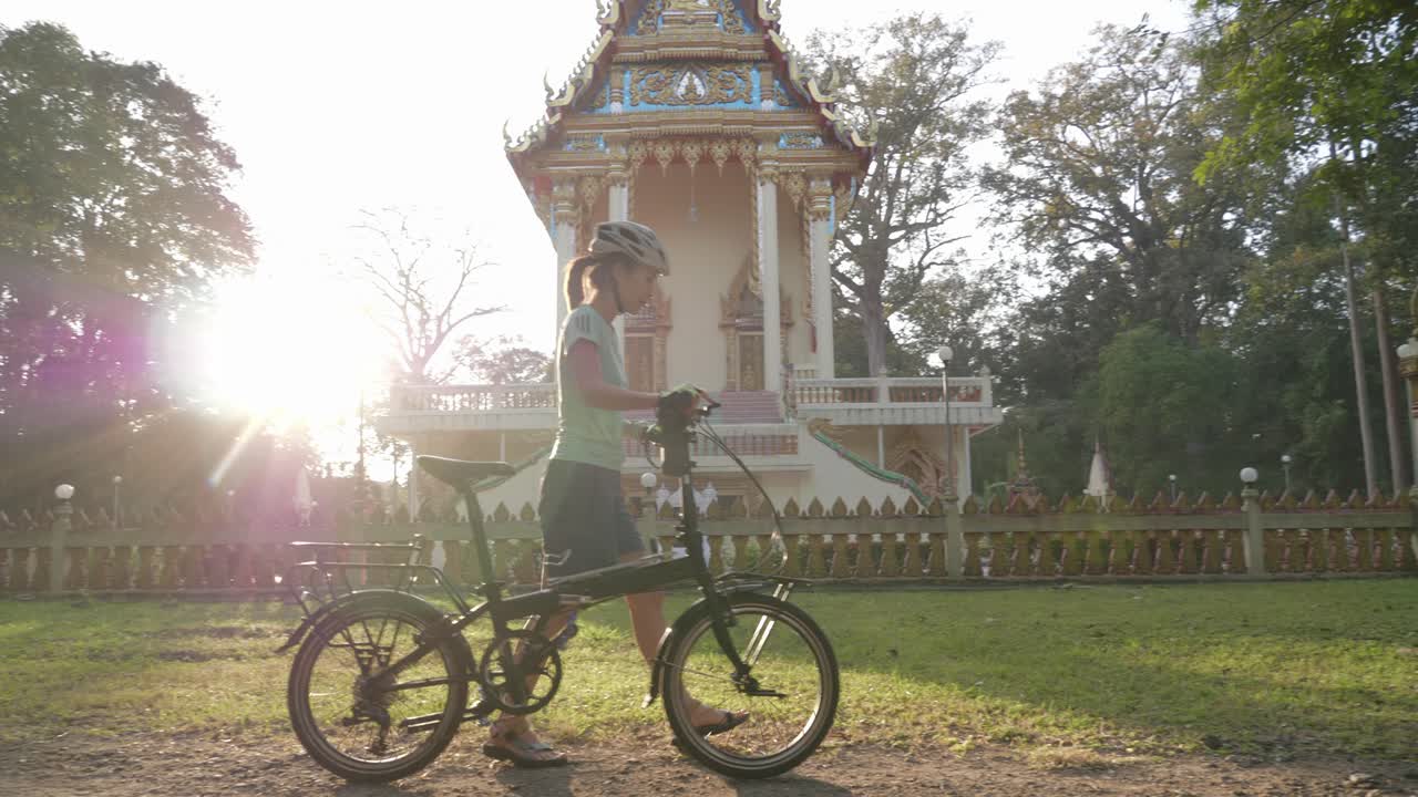 Una ripresa panoramica fissa di una ciclista che cammina con la sua bici passando accanto a un tempio sacro in Isan, Thailandia. Lei fissa il tempio mentre spinge la sua bici lungo il marciapiede attraverso il bagliore del sole
