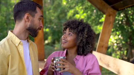 Young-black-woman-and-caucasian-man-have-a-cup,-talk-and-laugh-in-the-woods.-Pretty-female-with-afro-hair-outdoors.-Camping-day-in-the-mountains.