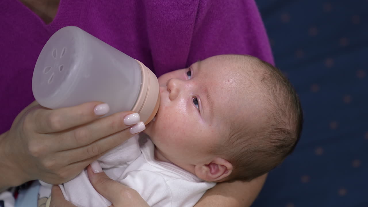 Sleepy baby finishing his milk in the bottle. Little child in mother's arms falling asleep gradually while having meal. Close up.