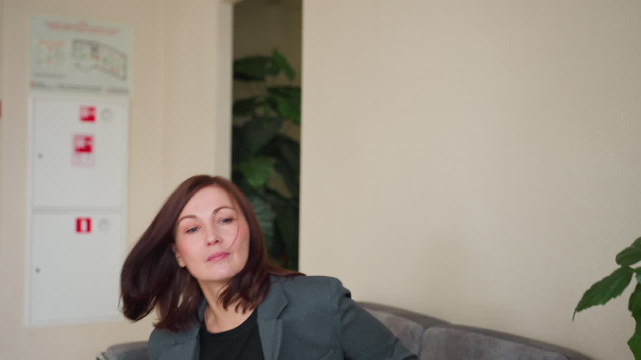 Confident businesswoman in gray blazer and black shirt standing in modern office hallway with serious facial expression and strong posture under Russian exit sign near staircase and indoor plants