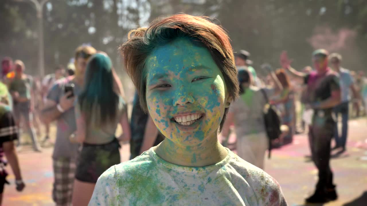 Young happy asian girl is smiling with colourful powder on holi festival in daytime in summer, watching at camera, color concept
