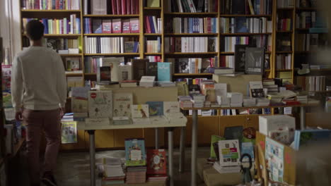 Young-man-looks-around-the-book-store-filled-with-different-books-on-the-display-table-and-book-shelves,-light-coming-in-the-room-from-the-left-side-thorugh-the-window