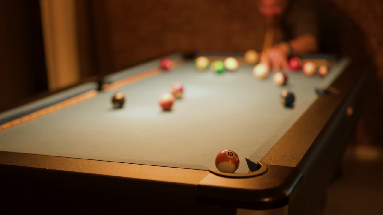 Man strikes cue ball on billiard table, low warm lighting, shallow focus, indoor leisure setting