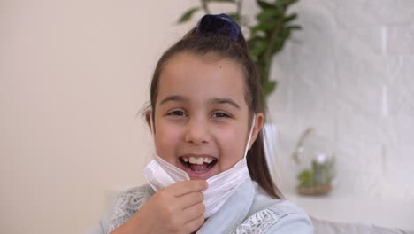 la niña se quita la máscara médica protectora y sonríe. quédate en casa mamá. coronavirus o covid-19. niña linda con una máscara protectora en casa en una habitación soleada y brillante