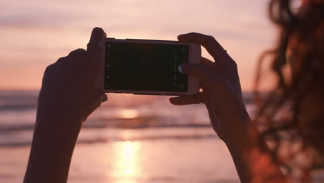 スマートフォンを使って美しい夕暮れの写真を撮り ⁇ ソーシャルメディアで夏休みの旅行体験を共有することを楽しんでいるビーチの若い女性