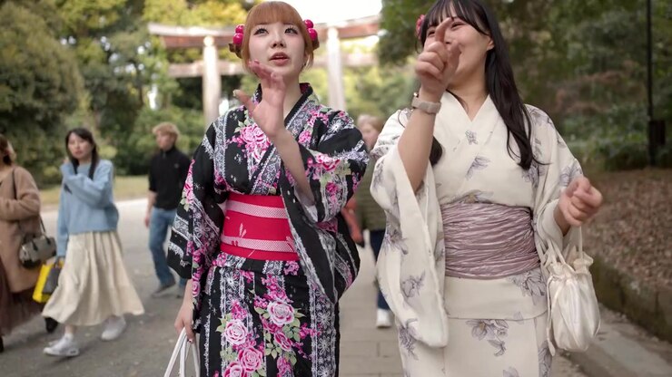 Due donne che indossano yukata tradizionali camminano e parlano attraverso un parco boscoso di un santuario shintoista.