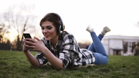 happy young female listening music on smartphone with headphones lying on green grass in park. girl having fun using her mobile phone outdoors