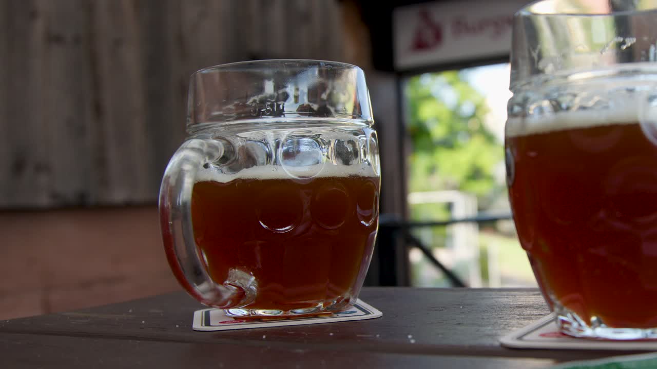 Glass beer mug on wooden table, hand reaching, sunny outdoor beer garden, natural daylight
