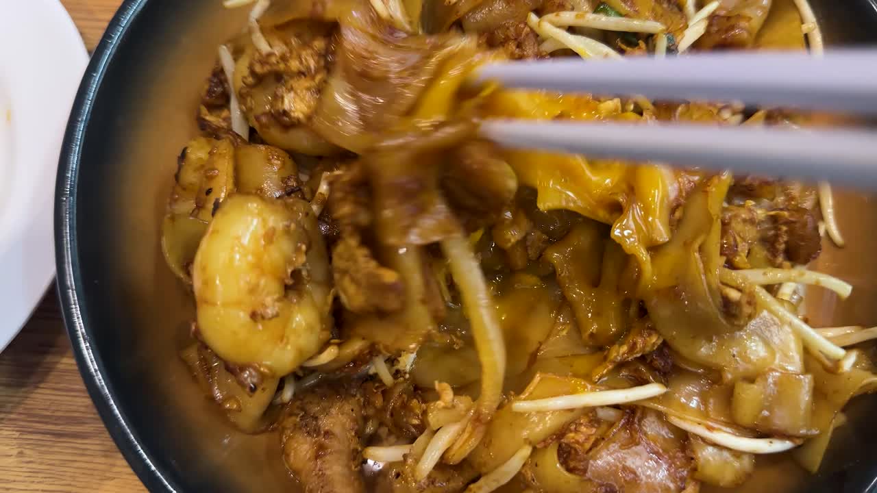 Chopsticks lift stir-fried shrimp noodles in bright lighting, overhead view, wooden table background