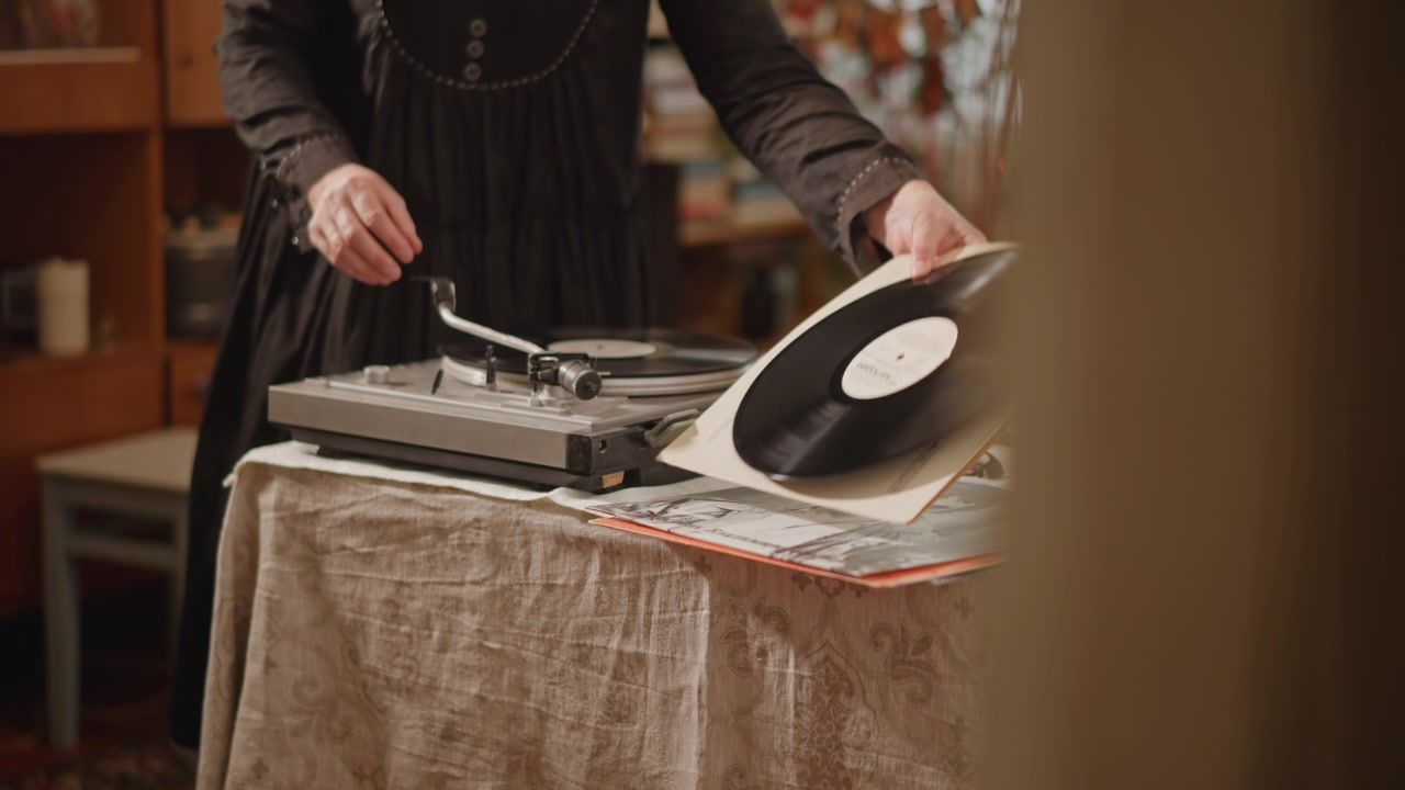Mani che posizionano un vinile sul giradischi, collezionista che sistema dischi su un tavolo coperto di stoffa con decorazioni vintage calde, candele e libri nelle vicinanze, posizionamento attento dell'ago e movimento lento tattile, nostalgico.