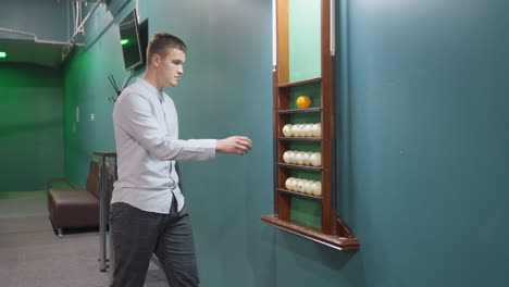 hombre con camisa blanca y pantalones grises entrando en la sala de billar hacia el rack de bolas de billar. paredes verdes, mesa de billar, iluminación aérea y interior elegante crean una atmósfera de juego relajada