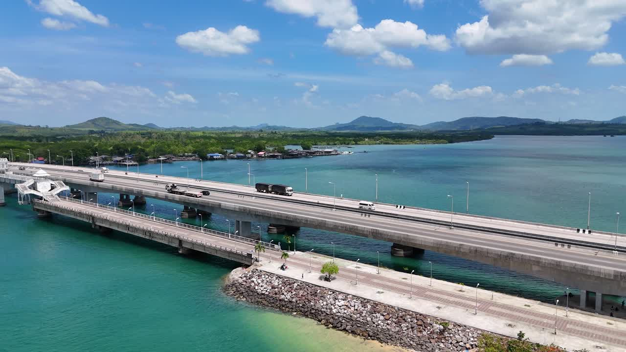 Drone footage captures Sarasin Bridge in Phuket, Thailand. Clear skies, turquoise waters, and moving vehicles create a serene, dynamic scene