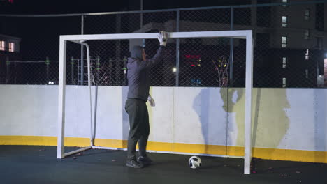 athlete walking towards soccer ball on ground while holding goal post during night training on urban outdoor sports court, background includes metallic fence and shadows