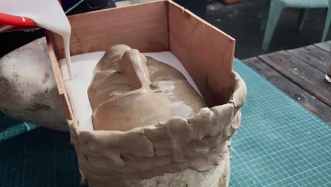 artista elaborando un molde facial mediante la fundición delicada de una mezcla de yeso líquido en un busto de retrato enmarcado en madera - primer plano con la mano