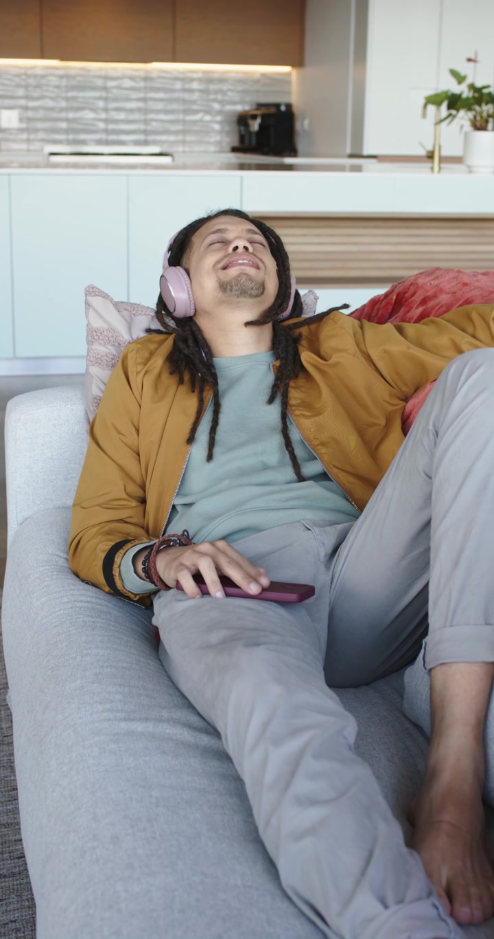 Video verticale: Uomo che entra in soggiorno, si sistema sul divano e indossa le cuffie per ascoltare musica rilassante.