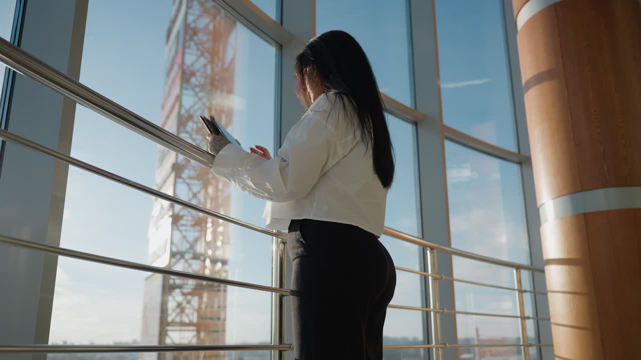 Rear view of young woman standing by metal railing holding tablet, interacting with device while overlooking cityscape through large glass window under clear daylight sky in modern interior