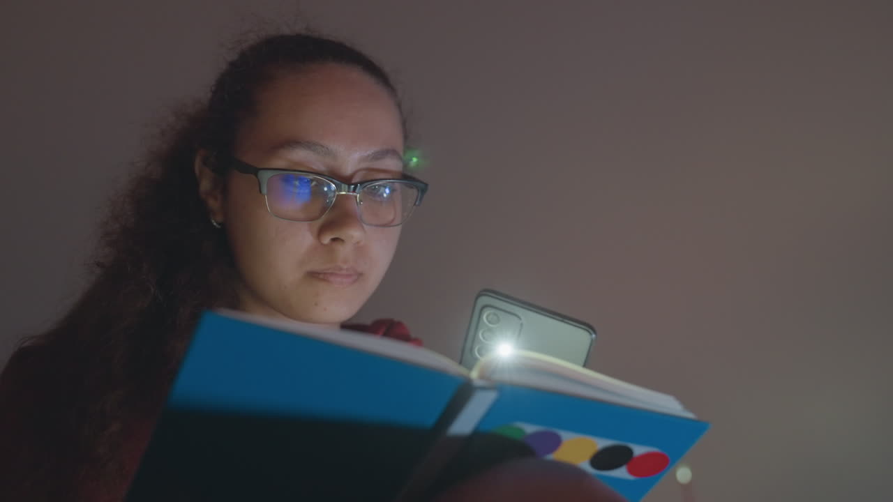 Young woman reading by phone light, latenight student studying notebook in dim bedroom, wearing glasses and curly hair, colorful dotted cover visible, phone glow illuminating pages, quiet concentrated