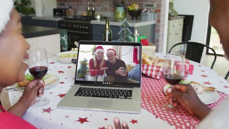 African-american-couple-with-wine-using-laptop-for-christmas-video-call-with-happy-couple-on-screen