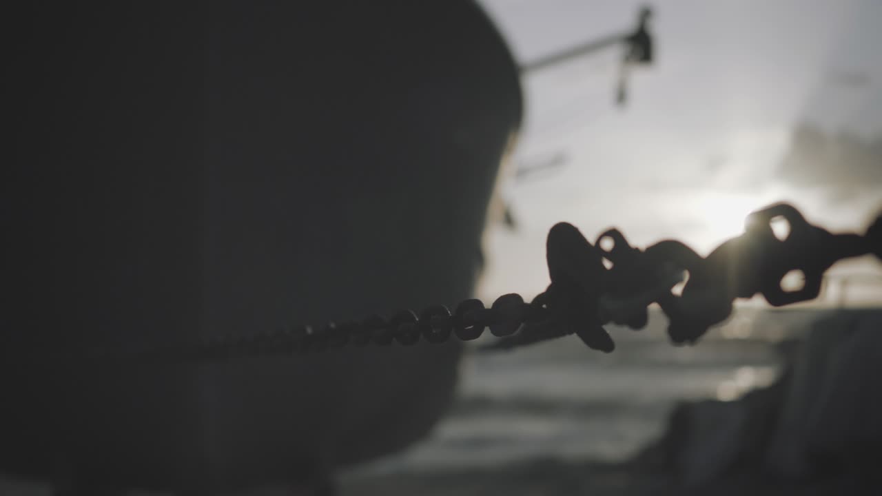 PARALLAX shot of a boat's chain and a LENS FLARE with the sun setting in the sea in the background.