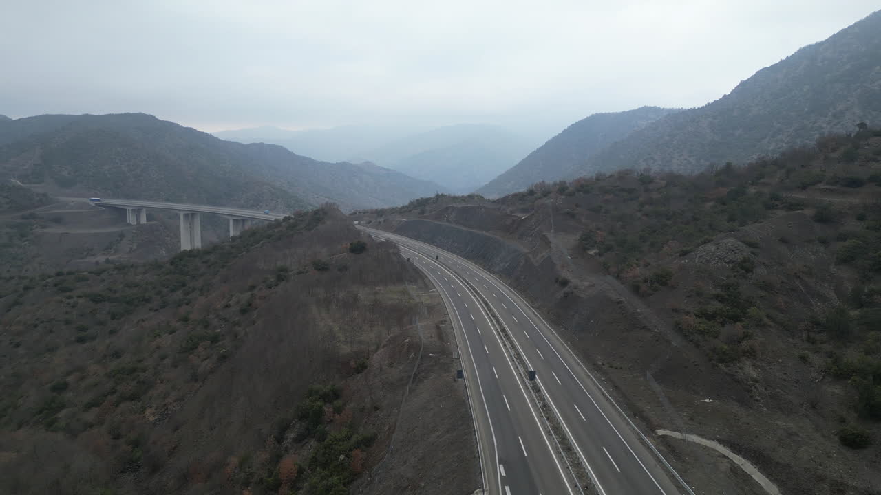 A drone soars along a Macedonian highway nestled between mountains. The sky is overcast, casting a serene, somber tone over the landscape, making the journey quietly captivating.