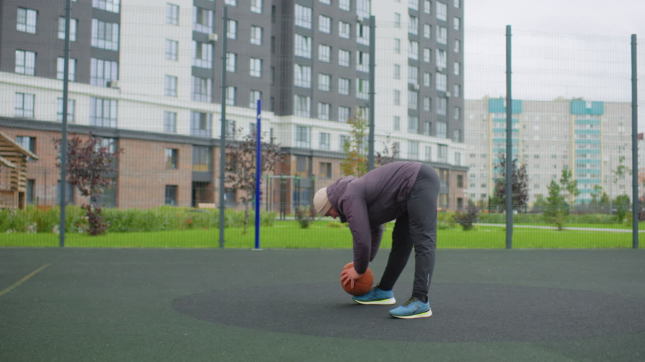 Senior white man dribbling basketball outdoors, balancing on skateboard, wearing puffer jacket and beanie, urban court beside apartment buildings, focused practice with controlled posture and steady