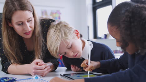 Female-school-teacher-sits-with-two-kids-using-tablet-computer-in-a-primary-school-class,-close-up