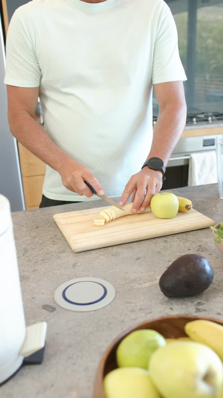 Video verticale: Posizionando una banana su un tagliere, l'uomo taglia a rondelle usando un coltello al bancone, preparando uno spuntino.