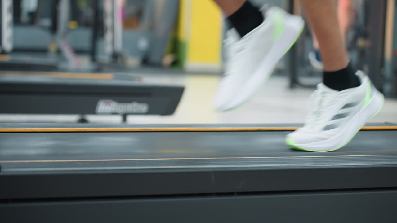 close up leg view of athlete running on treadmill with white sneakers, black shorts, dynamic motion blur, gym equipment and another person working out in background