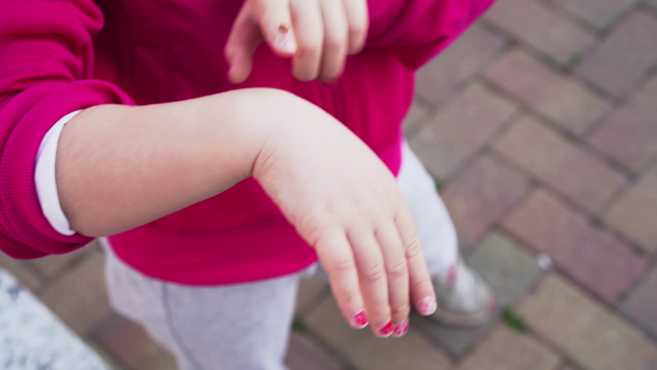 mariquita encima de la mano de una niña pequeña con jersey rojo y esmalte de uñas rosa sube a su dedo 4k