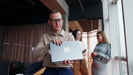 thoughtful male employee closes laptop and looks pensively into window. women at backdrop discussing work issues.
