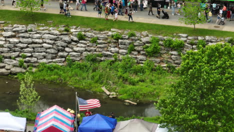 Colorful-tents-and-American-flag-at-Dogwood-Fest,-Arkansas,-USA,-near-a-creek-with-lush-greenery