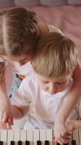 girl with braided hair plays melody on piano with brother fingers in children room upper view. sister teaches musical notes with blond toddler boy at home
