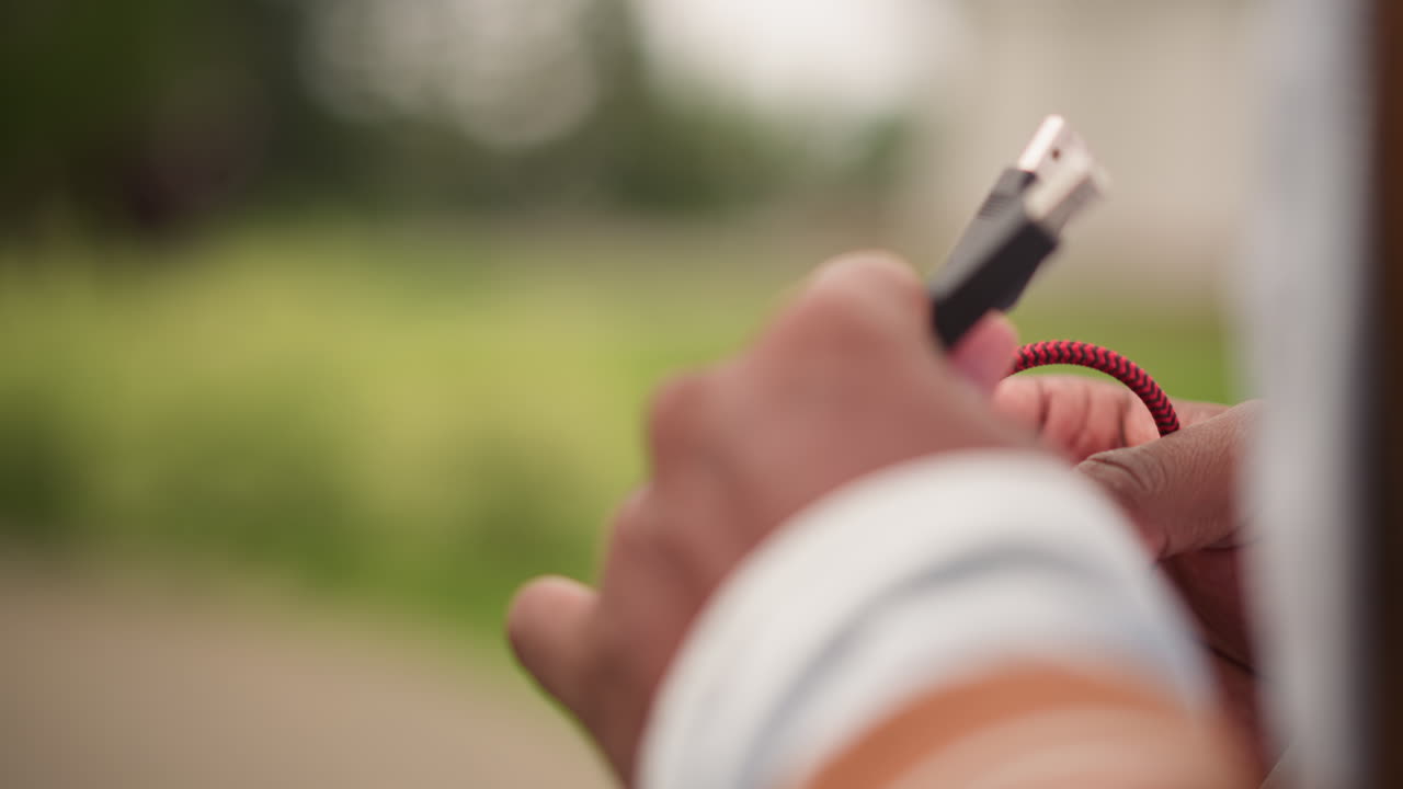 hands connecting audio cable outside near controller soft green background intimate focus on fingers and red coiled cable calm rehearsal mood natural light and casual jacket sleeve