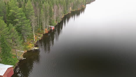 Sweden---Mirror-Lake-in-Autumn-Showcases-a-Colorful-Forest-on-a-Cloudy-Day,-Complemented-by-Red-Fishermen's-Houses-Along-the-Water's-Edge---Drone-Flying-Forward