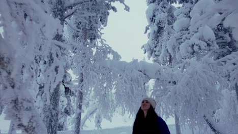 girl looking up under trees in snowy forest in snowy winter wonderland in lapland, finland, arctic circle