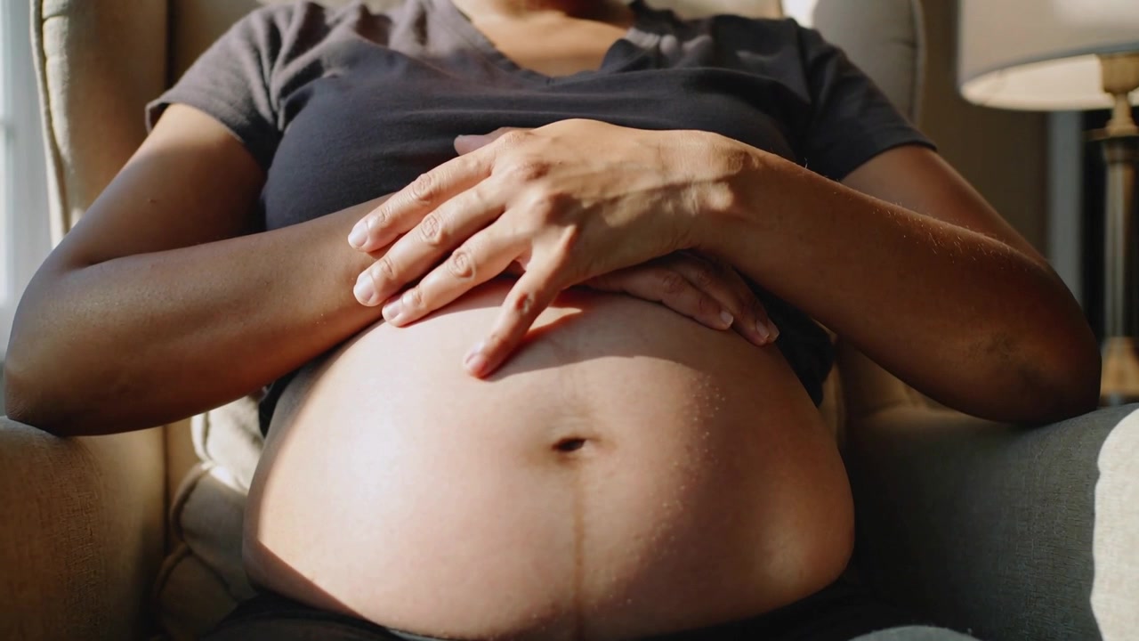 Hand lowering as pregnant woman tracing belly navel while sitting in armchair near window, dark tee