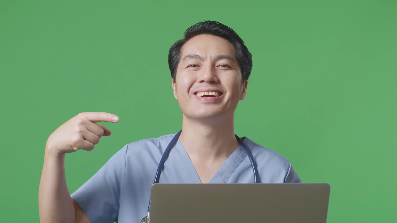 스테토스코프를 들고 있는 아시아 남성 의사가 웃으며 손에 노트북을 가리키며 병원에서 녹색 화면 배경에 서 있습니다.