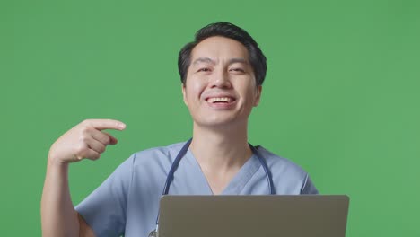 스테토스코프를 들고 있는 아시아 남성 의사가 웃으며 손에 노트북을 가리키며 병원에서 녹색 화면 배경에 서 있습니다.