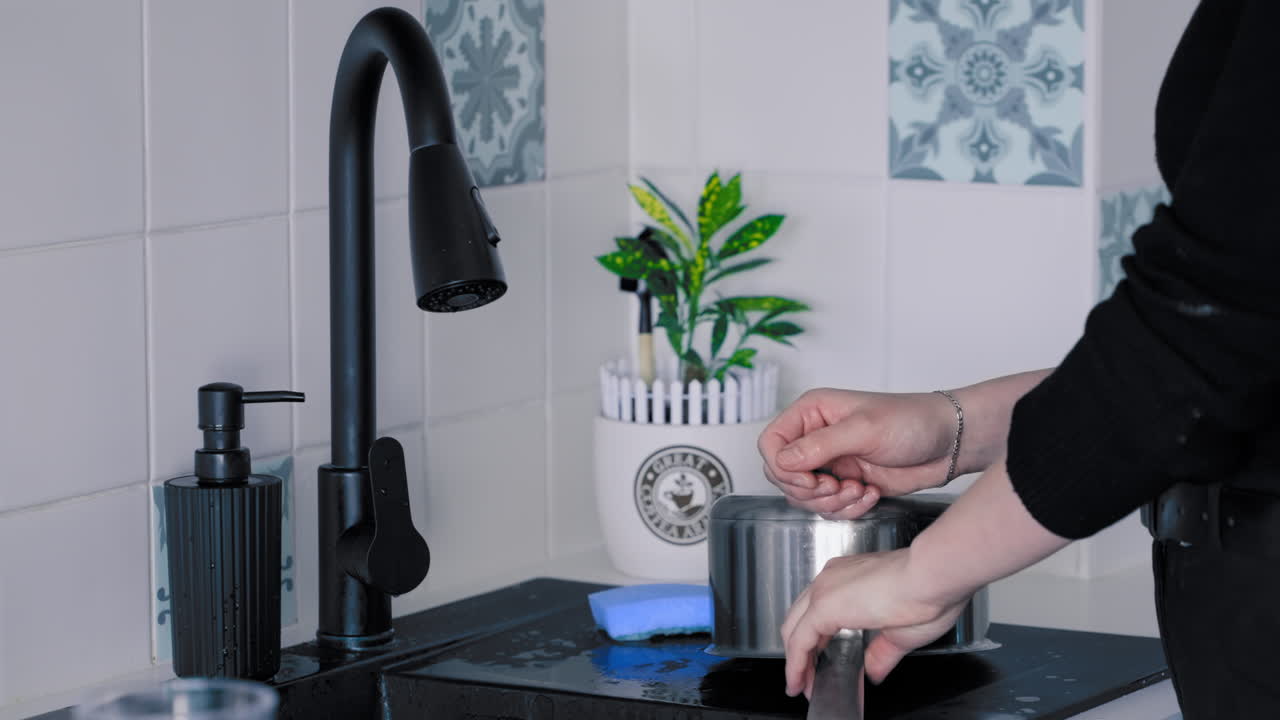 Primo piano di una giovane donna vestita di nero che lava i piatti con il sapone nel lavello nero della cucina sotto l'acqua corrente, al rallentatore.