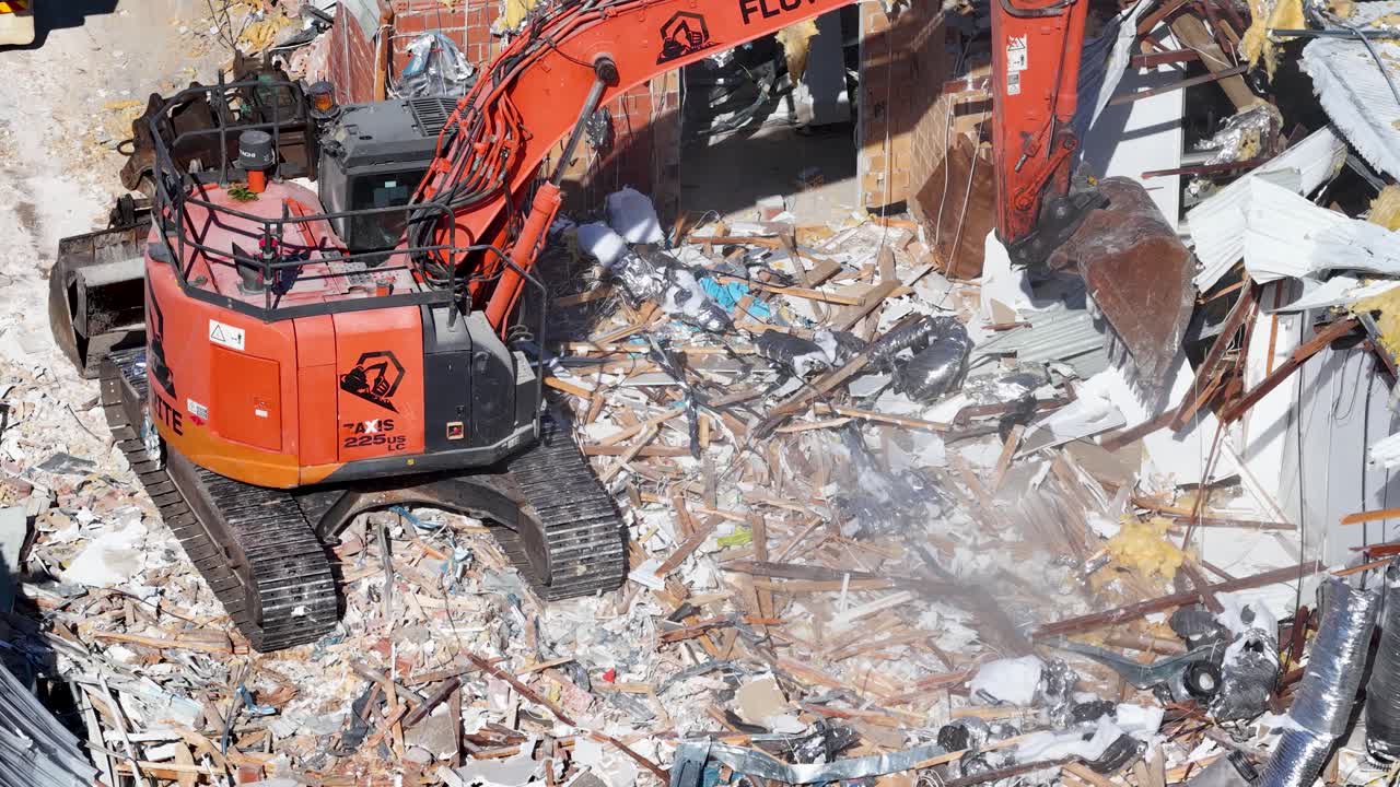 Large orange excavator tears down residential structure amid debris in bright daylight, with camera capturing steady aerial view of demolition process
