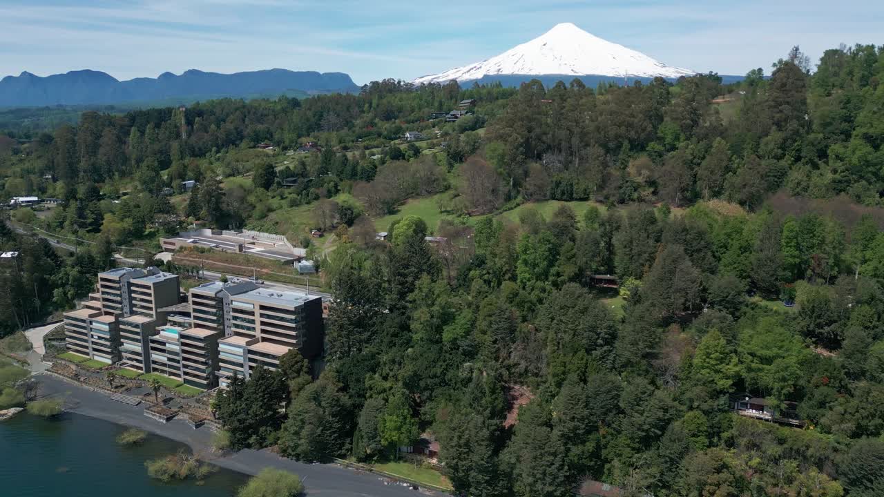 Aerial View of Lakeside Buildings Surrounded by Forest With Snow-Capped Volcano in the Background on a Clear Day
