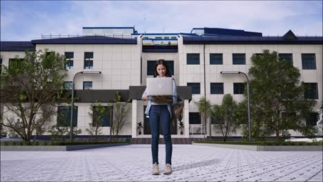 full body of asian teen girl student with a backpack smiling and typing on a laptop while standing in front of a school building