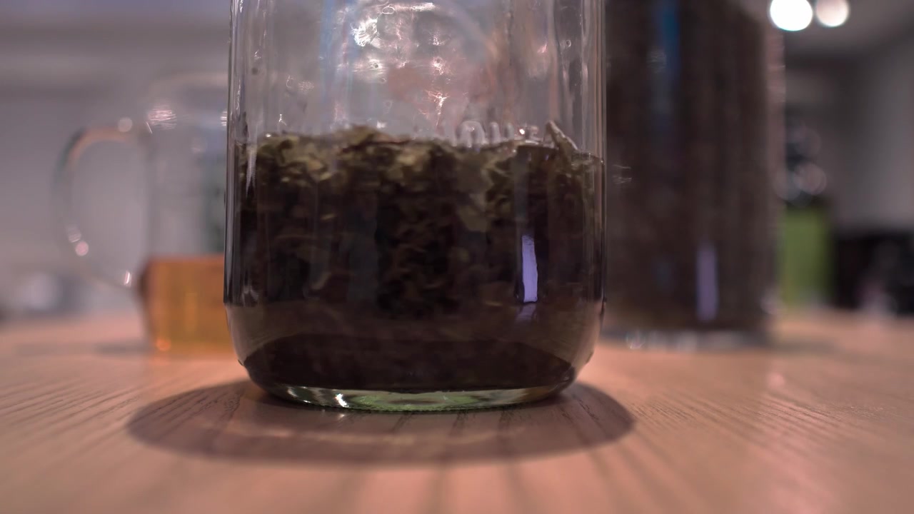 Close-up shot of a glass jar with loose tea leaves in liquid being placed on a wooden kitchen table next to a measuring glass and tea container.