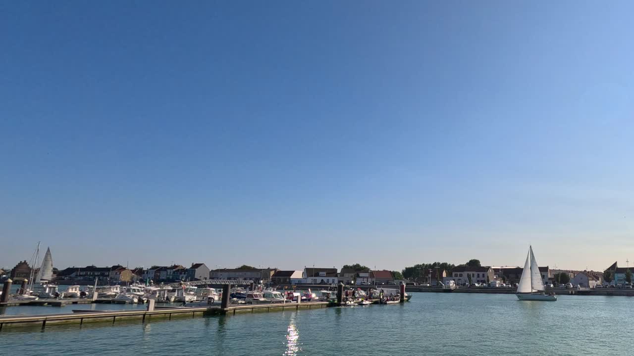 White sailboat moves across calm marina waters, clear blue sky, steady wide shot, natural lighting