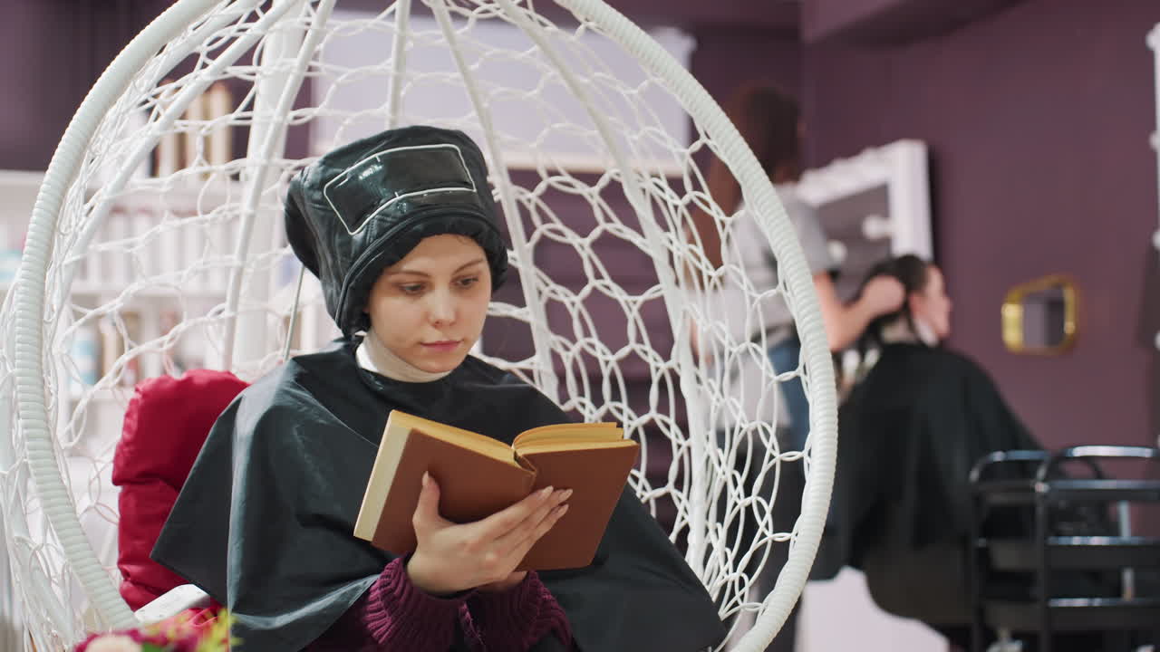 미용실에서 책을 읽는 여성, 보호 망토와 보닛 드라이어를 착용한 차분한 고객이 흰색 등나무 행잉 의자에 앉아 흐릿한 스타일리스트의 활동과 따뜻한 보라색 실내 조명 속에서 소설에 집중하고 있어요.