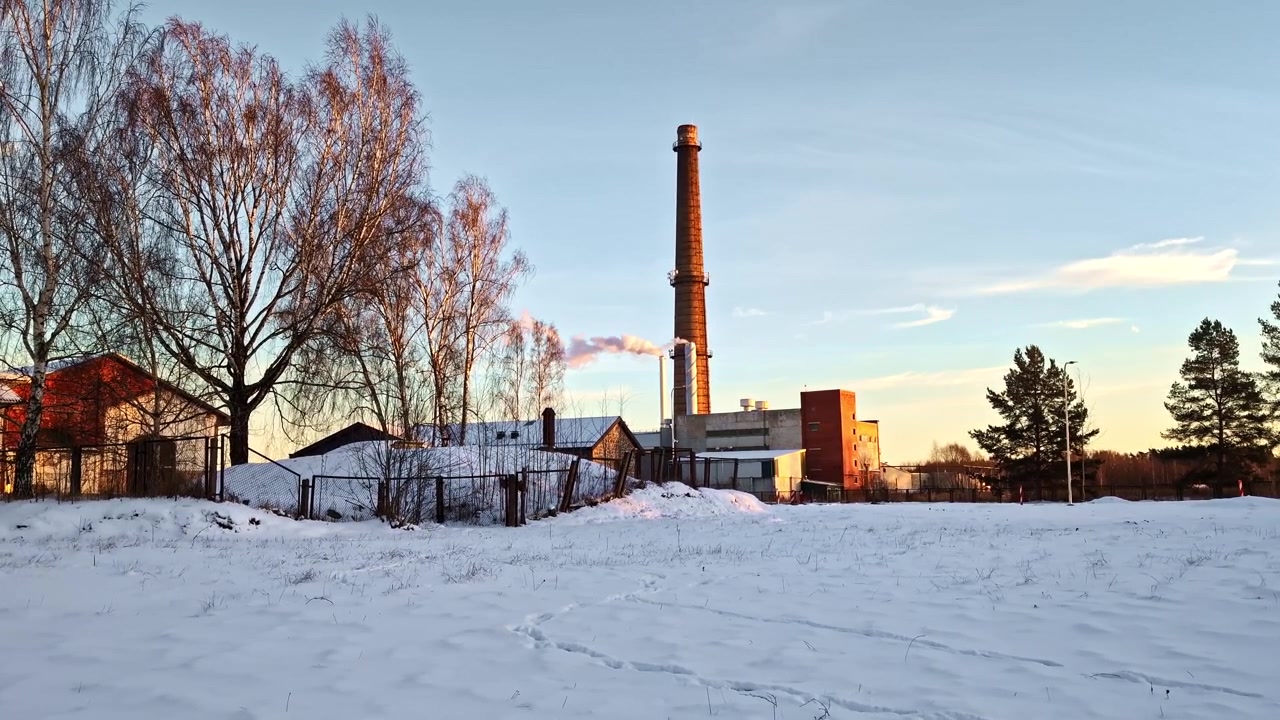 Industriel murstensskorsten, der udleder røg i et landligt snedækket landskab under en smuk vinter solnedgang.