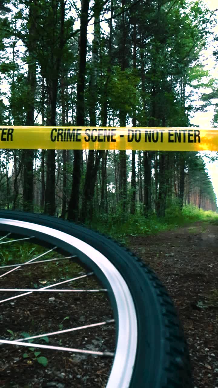 Eerie and scary video footage of a bicycle wheel spinning after an accident or crime while a yellow colored Crime scene Do Not Enter ribbon tape moves in the wind above it. Lonely mud road at a forest