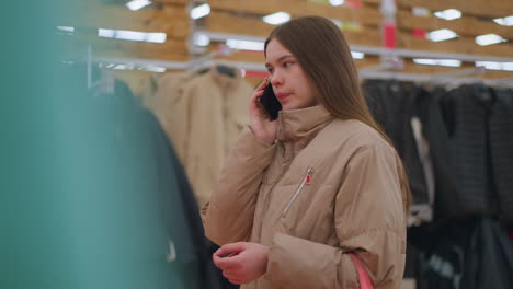 a young girl wearing a light brown jacket is standing in a mall, holding a phone to her ear as she engages in conversation. she appears to be holding a bag in her other hand