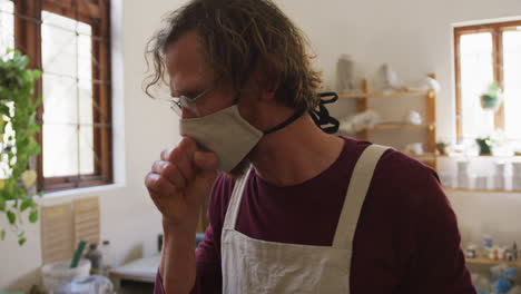 male caucasian potter wearing face mask and apron coughing at pottery studio